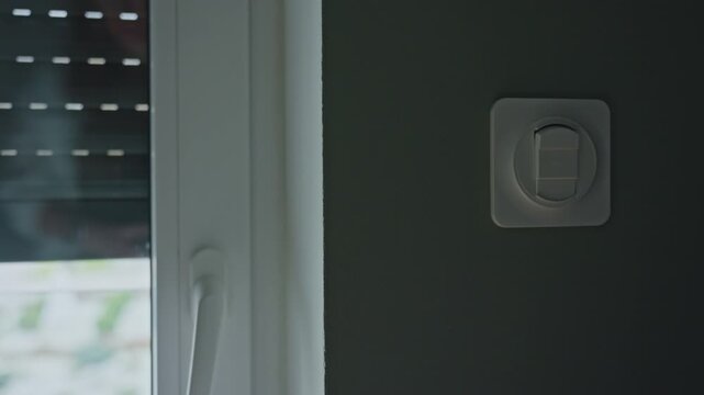 Woman pressing wall switch while exterior roller shutter goes down on window. Concept of home automation, light control and residential security