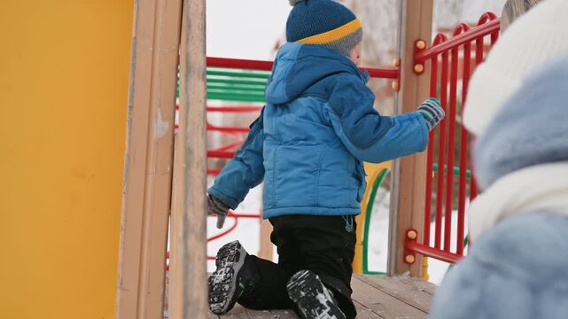 Young son climbing on snowy platform and crawling through play structure while mom watches in warm coat, blue puffer and pompom hat, apartment buildings backdrop, independent exploration, confidence