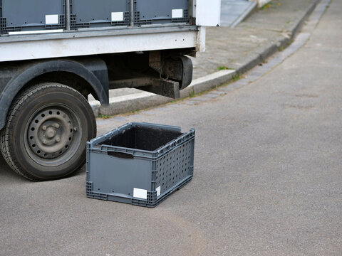 Close-up of a grey plastic Eurobox container sitting on the asphalt street
next to a white delivery truck. The scene shows logistics and distribution
in an urban environment. More stackable crates 