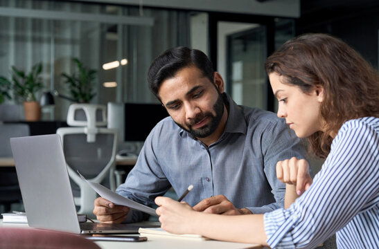 Two multiethnic professionals colleagues working together with laptop and papers in office. Indian male mentor and latin female young professional sitting in creative office space.