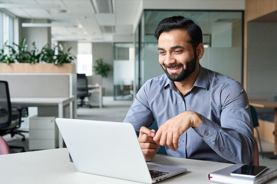 Happy smiling ethnical indian businessman having online virtual meeting call on laptop looking at screen sitting at table in coworking creative office, professional manager talking to employees.