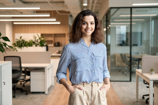Young smiling latin girl college student or teacher looking at camera standing in university campus. Happy hispanic millennial woman professional posing in modern coworking creative office space.