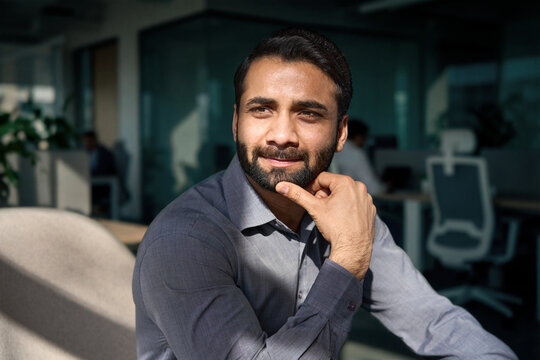 Portrait of young adult thoughtful bearded indian business man executive looking away sitting on chair in office lobby thinking of new ideas, planning project, dreaming of success lit with sunlight.