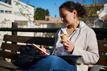 Obraz premium Woman Using Phone and Drinking Coffee While Working Remotely on a Bench Outdoors