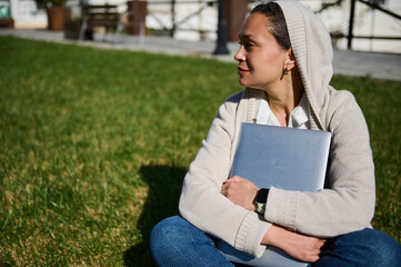 Fototapeta premium Young Woman Sitting on Grass With Laptop Outdoors, Thoughtful Remote Worker