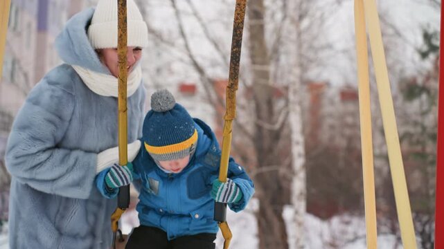 Mom pushing son on swing, joyful winter park scene with mother in blue coat and knit hat pushing toddler on classic swing seat, laughter on cold air, snowcovered ground, apartment buildings and bare