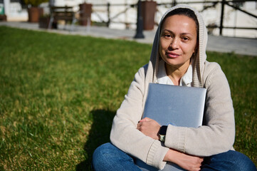 Fototapeta premium Relaxed Woman Sitting on Grass Outdoors Hugging Laptop - Casual Student Working in Park
