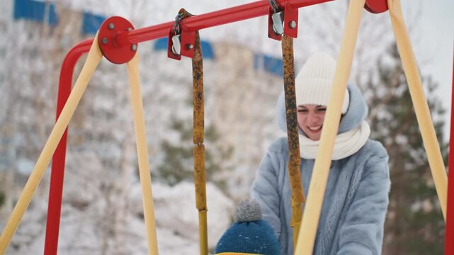 Mom pushes son on swing snowy playground laugh, child bundled in blue hat and puffer coat, mother wearing white knit hat and scarf, joyful interaction with visible breath, red swing frame, urban