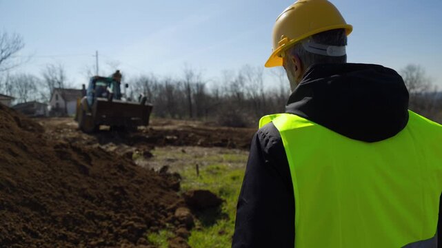 Wide shot of a heavy excavator machine moving dirt on a construction site while an engineer in a safety vest and hard hat observes the progress with project plans.
