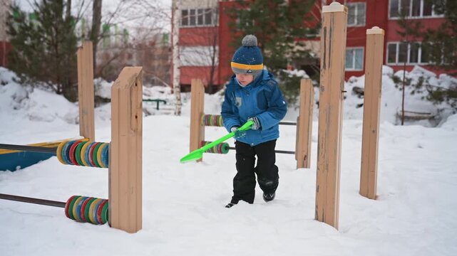 White son playing with green shovel bundled in blue jacket and beanie exploring urban playground digging in fresh snow curious expression wooden play structure footprints serene winter morning candid