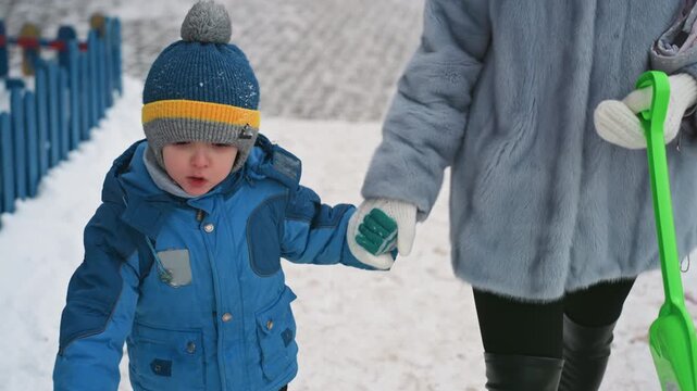 Mom and son walking in snow, holding hands beside blue fence, bundled in matching blue coats and knit beanie, mother guiding child with green shovel in hand, breath visible in cold air, footprints