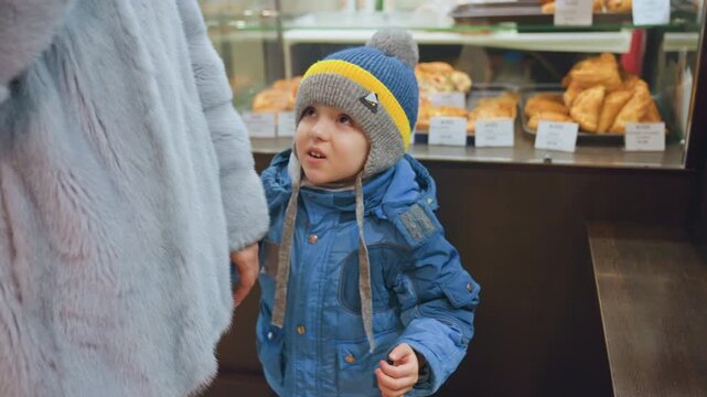 Mom and son at bakery counter, toddler in blue coat and knit hat reaches for pastry while mother in fur coat holds hand, croissant and bread display behind glass, warm indoor light, cozy morning