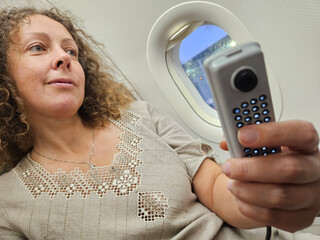 Woman is sitting on an airplane and holding a remote control. She is smiling and looking out the window © Pavel Losevsky
