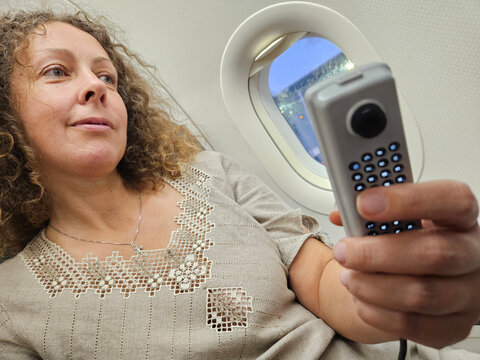 Woman is sitting on an airplane and holding a remote control. She is smiling and looking out the window