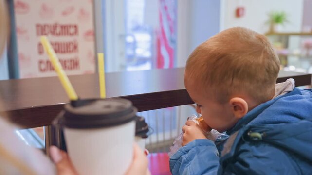 White mom and son at counter sharing pastry and warm coffee indoor cafe scene, mother holds takeaway cup while toddler in blue coat inspects straw and bites pastry, gentle interaction, cozy lighting,