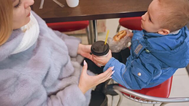Mom sharing snack with son at cafe table, donut and cup exchanged, child holding cup while mother lifts lid, playful interaction and laughter, close seating and cozy coat, gentle hand gestures, candid