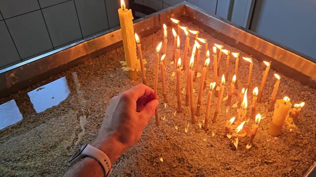 Person is holding a candle in front of a lit candle. The candles are arranged in a row and are lit