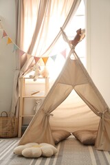Stylish toy wigwam, furniture, pillows and decor near window in playroom. Interior design © New Africa