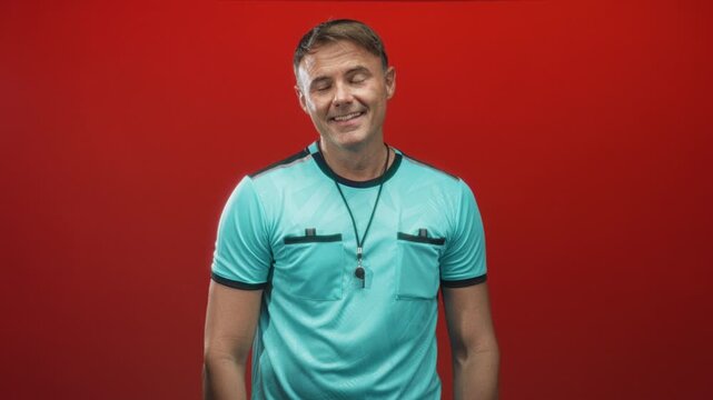 Man smiling with whistle on lanyard, wearing turquoise referee shirt against red studio backdrop; friendly officiating.