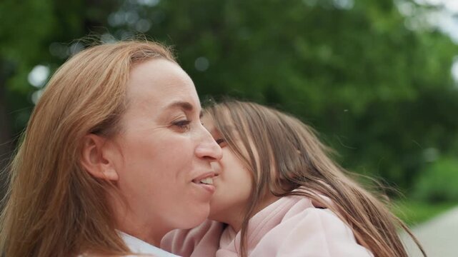 Mom daughter hugging outdoors park closeup soft kisses and laughter warm embrace mother holding child pink jacket green trees background candid joyful moment affectionate bonding nurturing connection