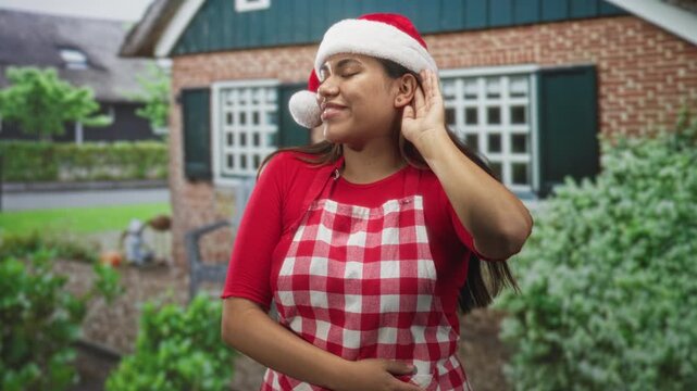 Woman in santa hat and checkered apron cups hand to ear outside a brick building while smiling and holding stomach; holiday joy.