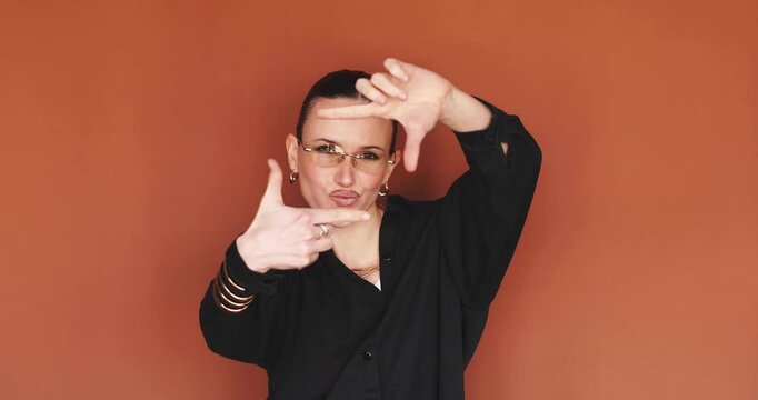 Cheerful woman in glasses making frame gesture with hands and looking at camera through fingers, playful female model posing with finger frame on orange background, focusing and creativity concept
