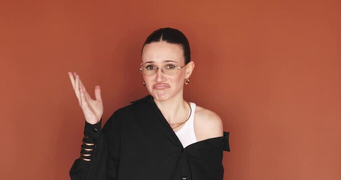 Displeased and skeptical young stylish woman with glasses showing stop and no gesture with hand, looking at camera with grimace of disgust and rejection, standing over terracotta brown background