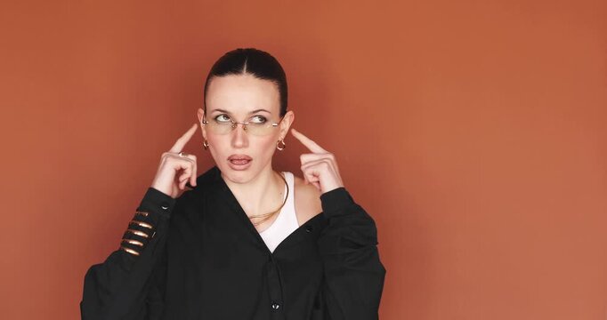 Annoyed young stylish woman covering ears with fingers not wanting to listen, looking away with irritation and ignoring loud noise or talk, standing over terracotta brown background in studio