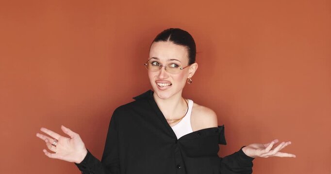 Embarrassed and sorry young stylish woman in glasses making oops face and covering mouth with hand, realized mistake and feeling regret, shrugging over terracotta brown background in studio