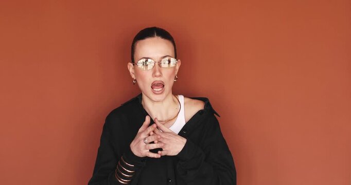 Portrait of a stylish young woman in yellow glasses looking at the camera with a confused expression and shrugging her shoulders asking why or what is happening on a solid brown background
