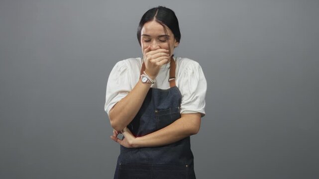 Young woman in apron covers mouth with hand in studio while stifling a laugh and crossing her other arm; surprise hospitality.