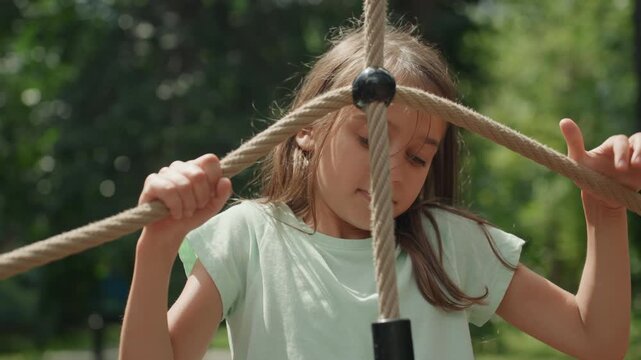 Focused daughter navigating rope web park, intent gaze through grid of ropes, careful leg placement on knotted crossings, denim shorts and white sneakers, sunlit green backdrop, concentrated play