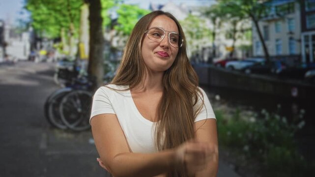 Woman with hand covering mouth and crossed arms on street by a canal and parked bicycles; shy amusement.