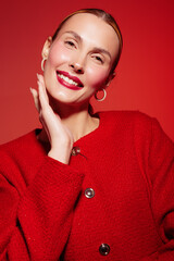 Smiling woman with red lipstick and hoop earrings poses in red sweater against red background. Close-up portrait with natural makeup and joyful expression. Concept of beauty and fashion. © SHOTPRIME STUDIO