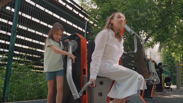 Outdoor exercise mom and daughter cooperating on machine, child pushing handle while mother steadies on seat, teamwork and laughter, urban greenery corridor, warm sunlight and tender family care