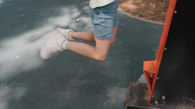 Young girl jumping from playground bar, midair legs bent, sneakers visible, smooth descent to rubber surface, summer park light dappled shade, denim shorts and casual tshirt, playful landing sequence,