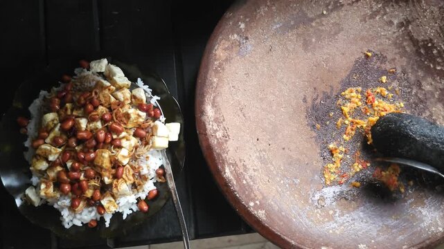 Preparing traditional Indonesian food by scraping freshly ground sambal from a stone mortar next to a dish of white rice with diced tofu and fried peanuts