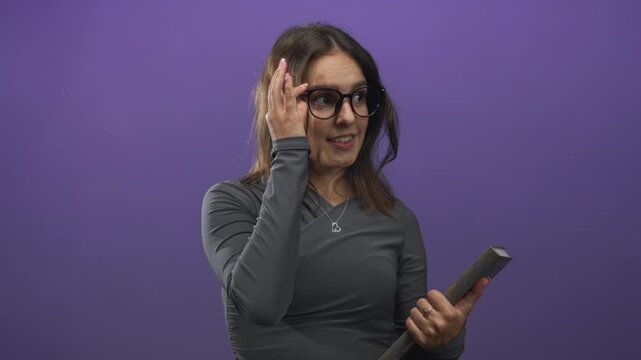 Young woman holding book and adjusting glasses while smiling in purple studio, wearing gray top and heart necklace; studious learning.