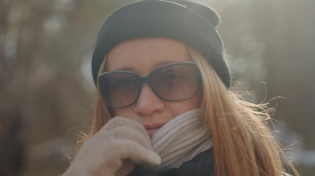 Smiling caucasian redhead in sunglasses with sunlit woods background. Beanie and scarf frame face, warm glow and lens flare add cinematic warmth. Closeup portrait shows confident grin and friendly