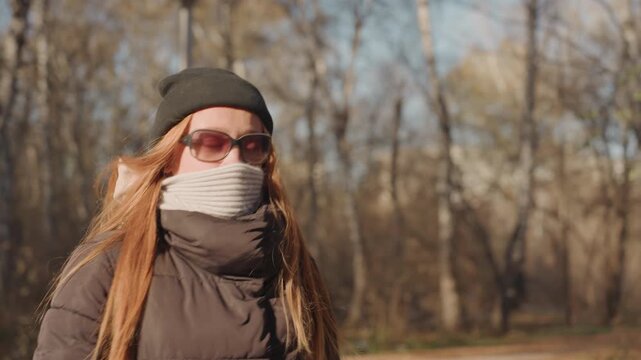 Caucasian woman frontal closeup walking through treelined avenue wearing sunglasses, mask and beanie, long red hair flowing and carrying a case backpack, assertive commuter stride with empty path