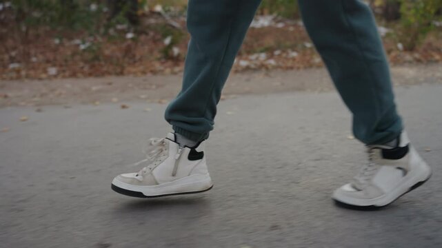 Person walking on paved park path, hoodieclad figure strides past trees and fallen leaves, gloved hand visible, closeup on sneaker soles on asphalt, subtle motion and overcast light create tense