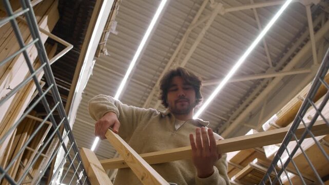 Low angle shot from shopping cart shows man loading purchased wooden beams, reaching forward to place long timber pieces into trolley under bright hardware store ceiling lights.