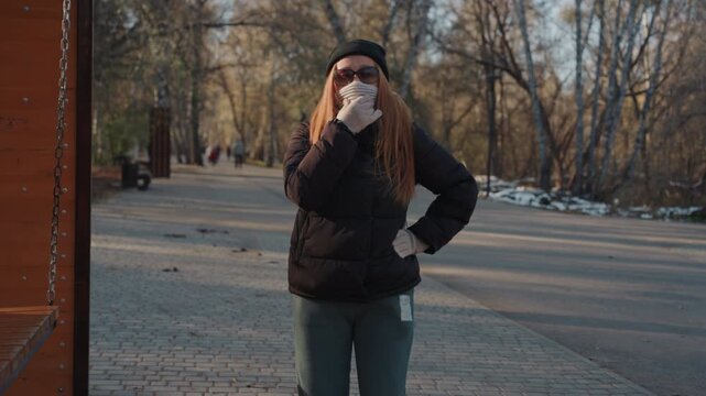 Woman in park covering face with scarf and sunglasses, bundled in dark coat and beanie, standing on empty autumn path, glancing around warily as if scouting for opportunity cinematic late sun