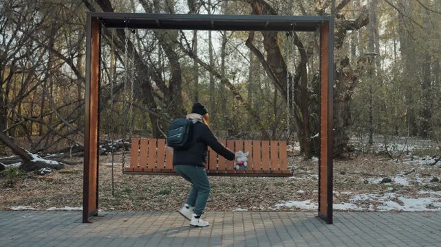 Running away from wooden bench, backpacked person sprints with hurried steps after snatch, rapid motion across snowy park path, tense chase energy, dusk light and bare trees intensify urgency.