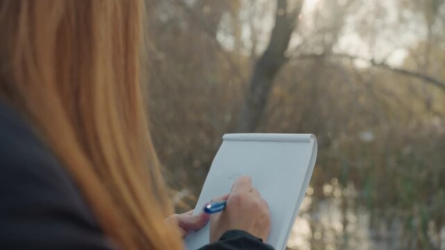 Woman in beanie writing pad outdoors, autumn river bank closeup of hands penning sketches and notes, somber light and muted colors, investigative tension as witness records prowler and snatcher