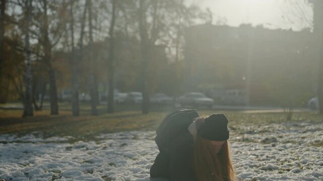 Backlit park white woman tosses snow during cold afternoon backpacked commuter pauses between errands, red hair haloed by sunflare, frosty grass and distant cars, soft bokeh, introspective mood
