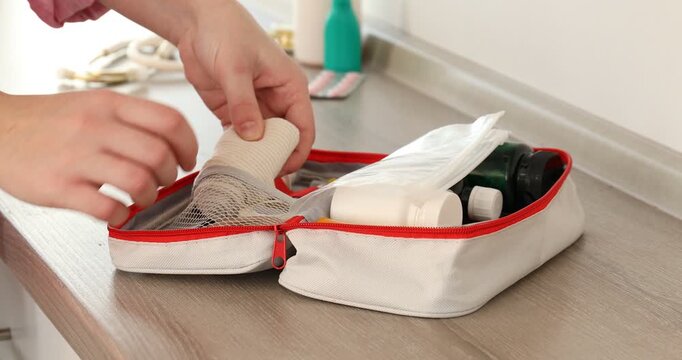 Woman putting elastic bandage roll into first aid kit at wooden counter near white wall indoors, closeup