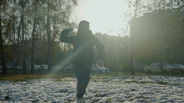 Backlit hooded figure walking across snow. Tense late afternoon light with lens flare, bare trees and parked cars in background, footprints on frozen grass, slow stride suggesting snatcher or prowler,