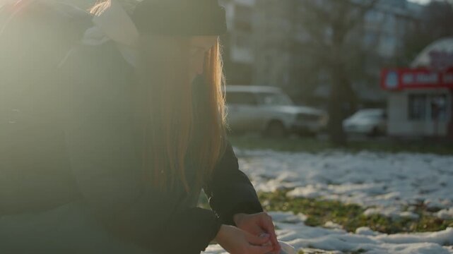 Woman crouching placing toy down in thawing snow by city street, backlit hands handle small doll with hesitant care, scene of possible snatcher returning or taking object, soft sunrise glow,