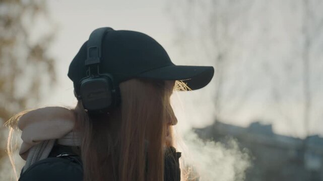 Caucasian girl with headphones in winter, cautious posture and visible breath, backlit city buildings, cap and scarf, listening to music while scanning surroundings for prowler and potential snatcher